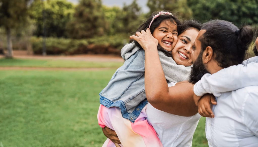 A mother hugs her daughter while the father looks at them both in a park.