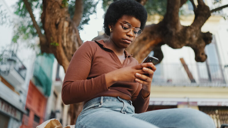 Smiling Family Including Grandparents Two Young Girls Pose Young Black woman with afro hairstyle and glasses looking at her phone outdoors.