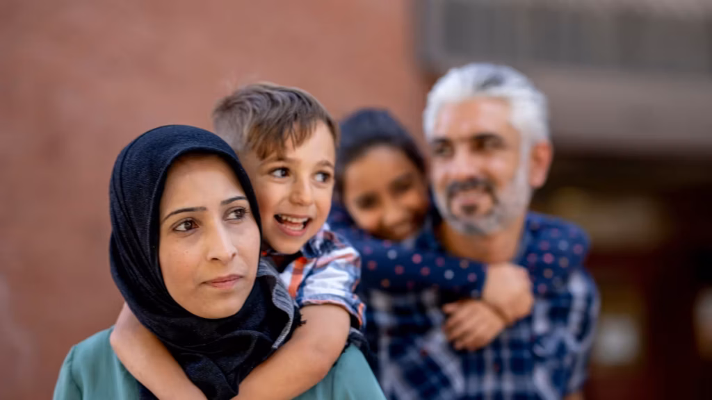 A mother wearing a hijab carries her son on her back while her husband and daughter are blurred in the background.