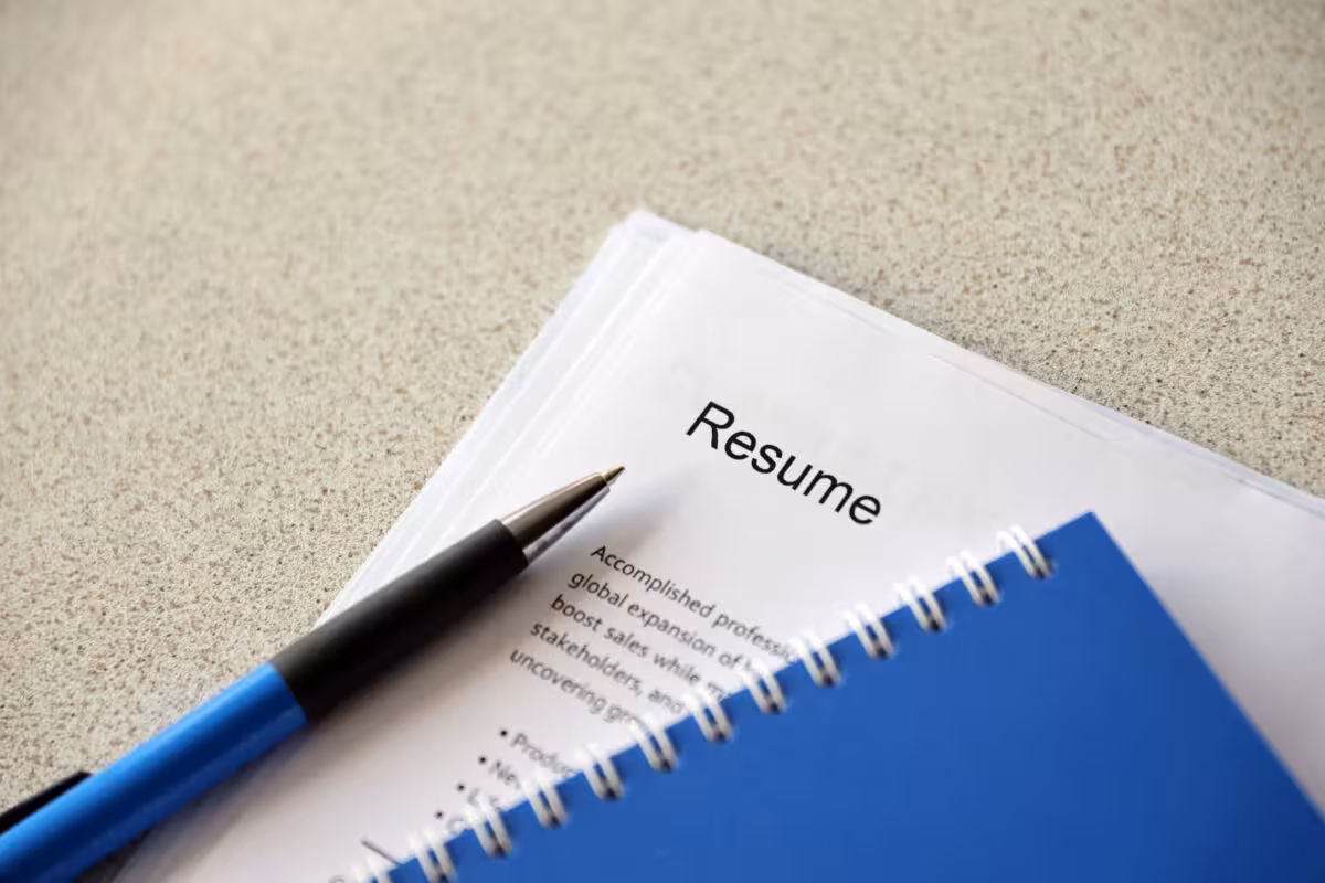 A pen rests on a resume next to a blue notebook on a textured surface.