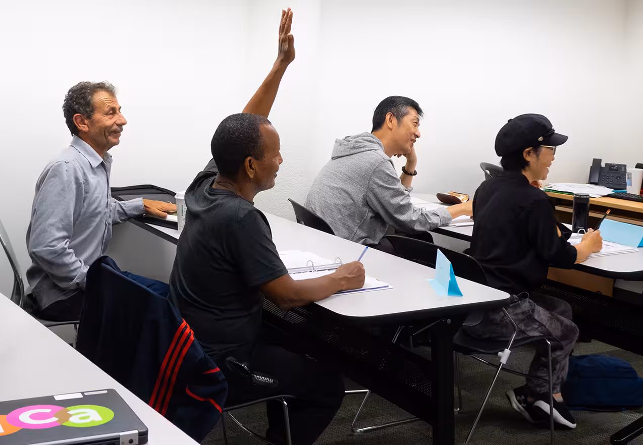 Four adults sit at tables in a classroom setting, with one man raising his hand.