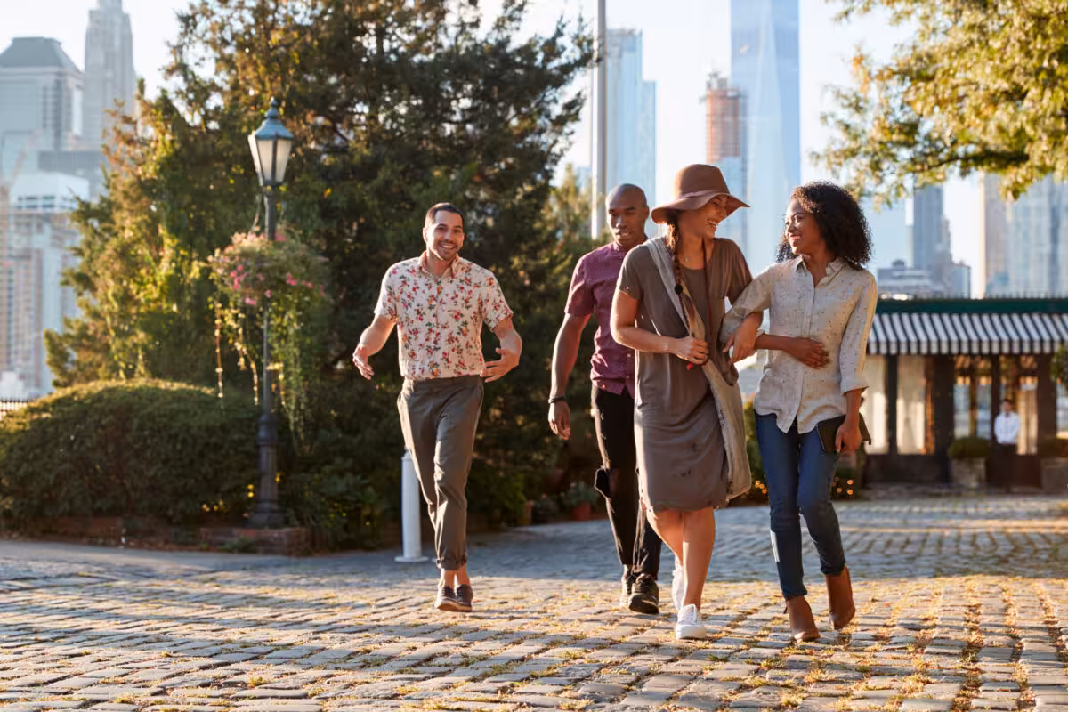 Diverse Group Four Friends Walks Along Cobblestone Path A diverse group of four friends walks along a cobblestone path in a city park, with buildings in the background.