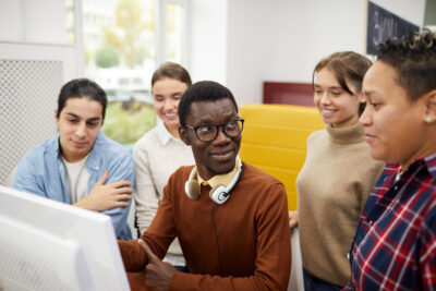 A multi-ethnic group of young adults collaborate around a computer monitor in a bright room.
