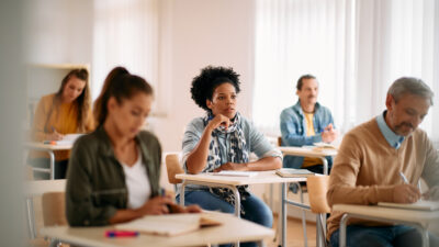 Diverse students attend class in a small but bright room, taking notes and listening attentively.