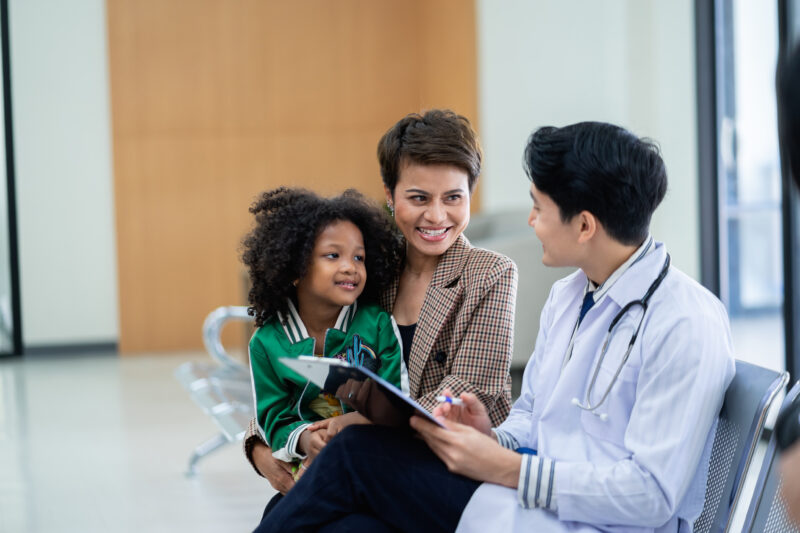 Doctor White Coat Stethoscope Talks Mother Her Young A doctor in a white coat and stethoscope talks with a mother and her young daughter in a waiting room.