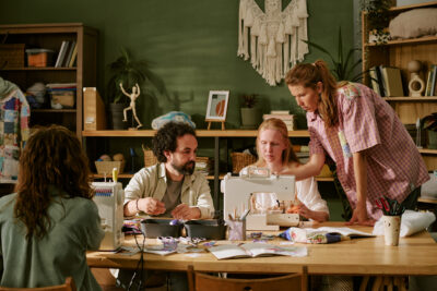 Four diverse adults work around a table with sewing machines, crafting in a well-lit room.