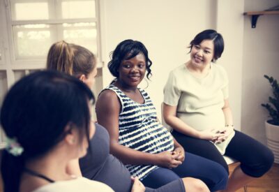 Four pregnant women of diverse ethnicities sit together, smiling and talking at a baby shower.
