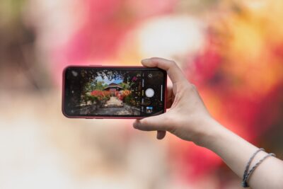 A light-skinned hand holds a smartphone horizontally, capturing a vibrant garden path lined with colorful flowers.