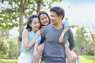 A happy girl enjoys a piggyback ride with her parents in a sunlit park with trees and a small tent.