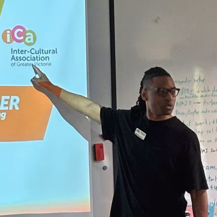 Man in black shirt and glasses points to a screen with the Inter-Cultural Association of Greater Victoria logo.