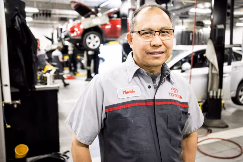 Man Wearing Toyota Mechanic Uniform Name Tag That A man wearing a Toyota mechanic uniform with a name tag that reads "Placido" smiles at the camera in a garage.