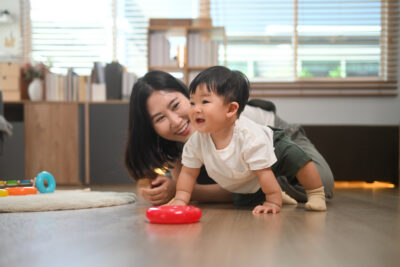 An Asian mother smiles as her baby crawls on the floor with toys nearby.