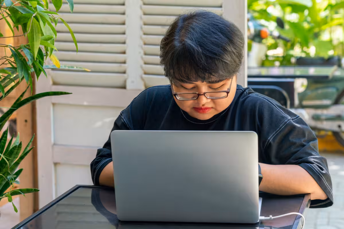 Person Wearing Glasses Looks Down Silver Laptop Computer A person wearing glasses looks down at a silver laptop computer on a table.