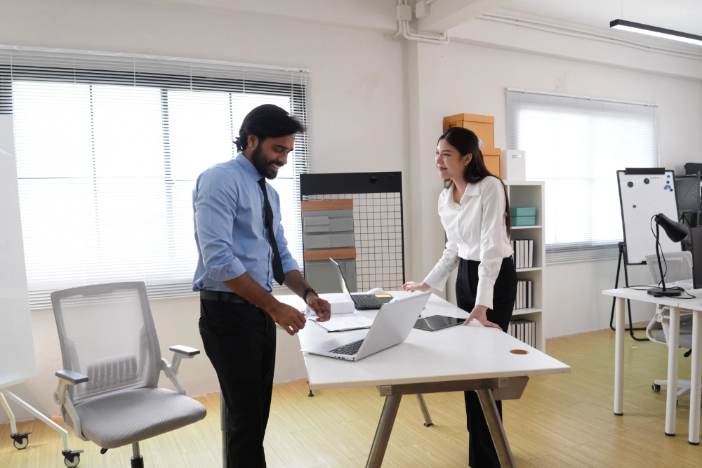 professional-man-and-woman-collaborating-at-a-desk-2026-10-02-00-27-34-utc A smiling woman in a hijab holds a smartphone while a diverse group of colleagues works in the background.