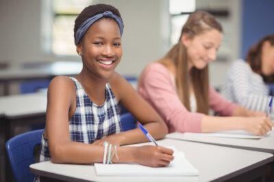A smiling Black girl in a plaid dress and headband studies at a desk in a classroom.