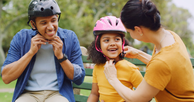 A smiling family puts on bicycle helmets in a park setting, preparing for a ride.
