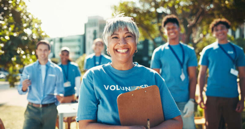 Smiling Woman Blue Volunteer Tshirt Holds Clipboard Diverse A smiling woman in a blue "VOLUNTEER" t-shirt holds a clipboard, with a diverse group of volunteers behind her.