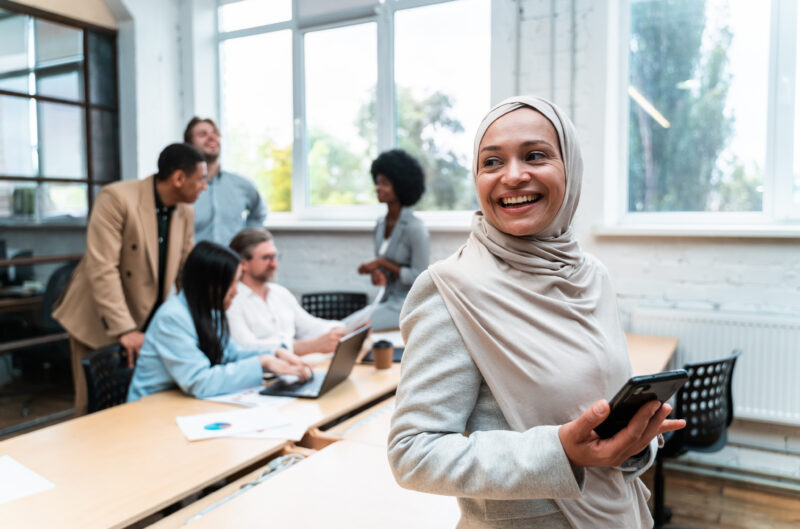 Smiling Woman Hijab Holds Smartphone While Diverse Group A smiling woman in a hijab holds a smartphone while a diverse group of colleagues works in the background.