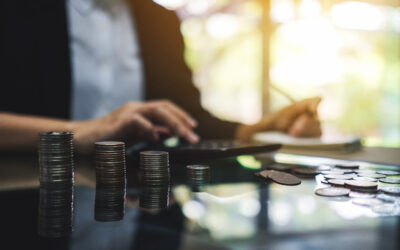 Stacks of coins and loose coins on a reflective surface with a person calculating in the background.