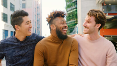 Three diverse men laugh together outdoors in a city setting with buildings in the background.