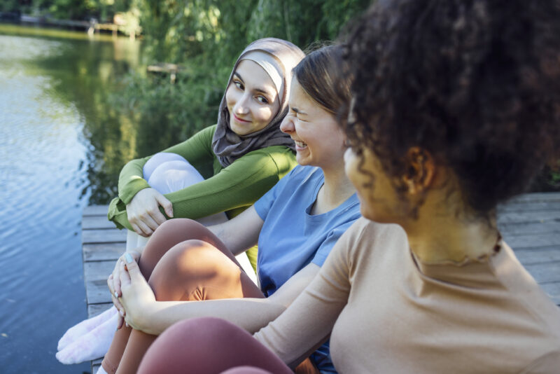 Three multiracial teenage women laughing while sitting on a dock by the water.
