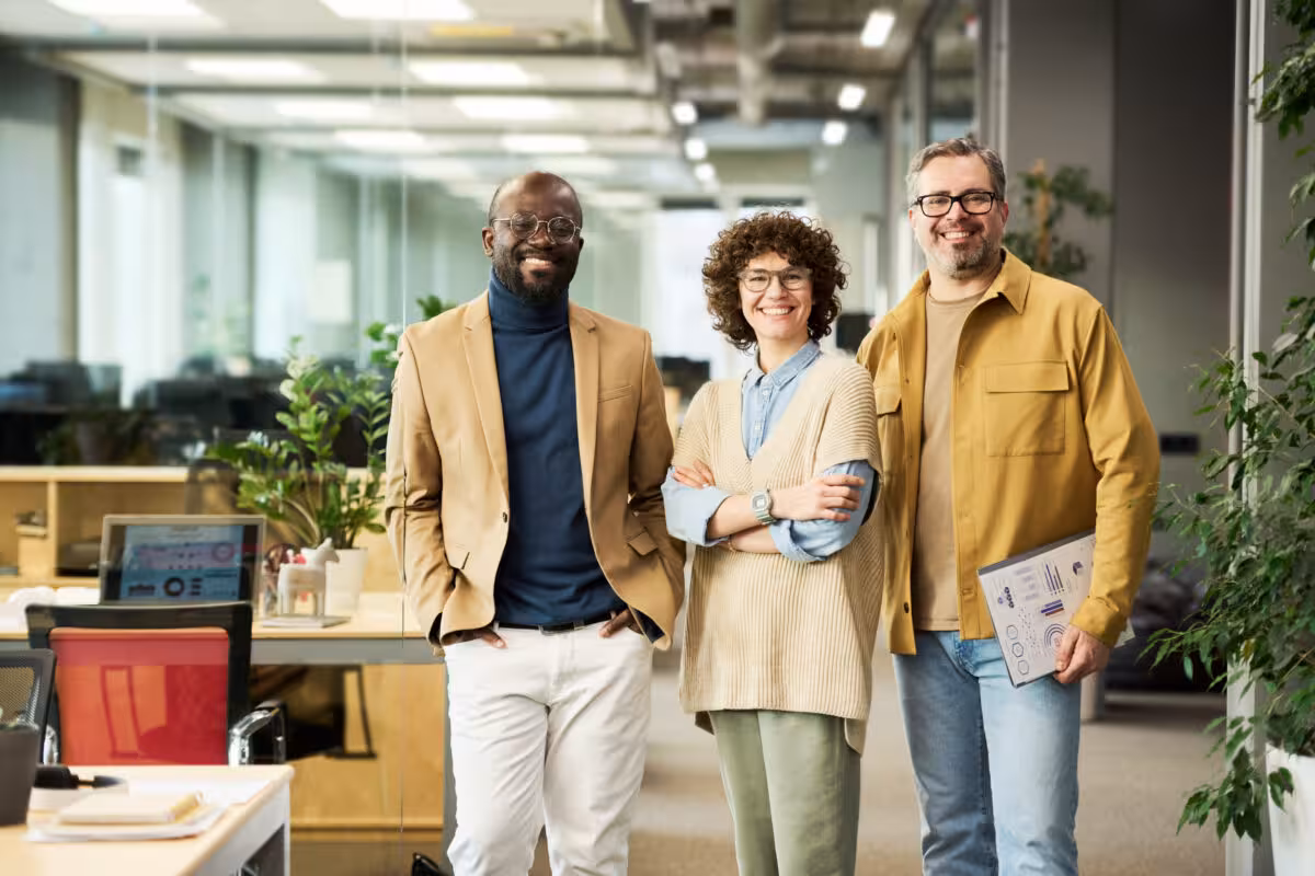 Three Smiling Colleagues Two Men One Woman Stand Three smiling colleagues, two men and one woman, stand in a modern office hallway with plants and desks.