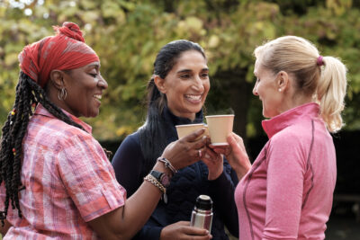 Three smiling women in their 50's enjoy coffee outdoors, toasting cups and holding a thermos.