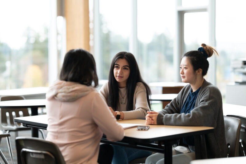 Three young women sit at a table in a classroom, talking to each other.