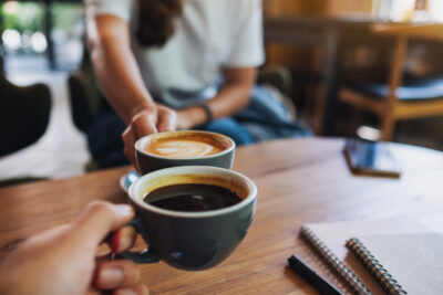 Two hands hold cups of coffee at a cafe table, with a notebook and phone nearby.