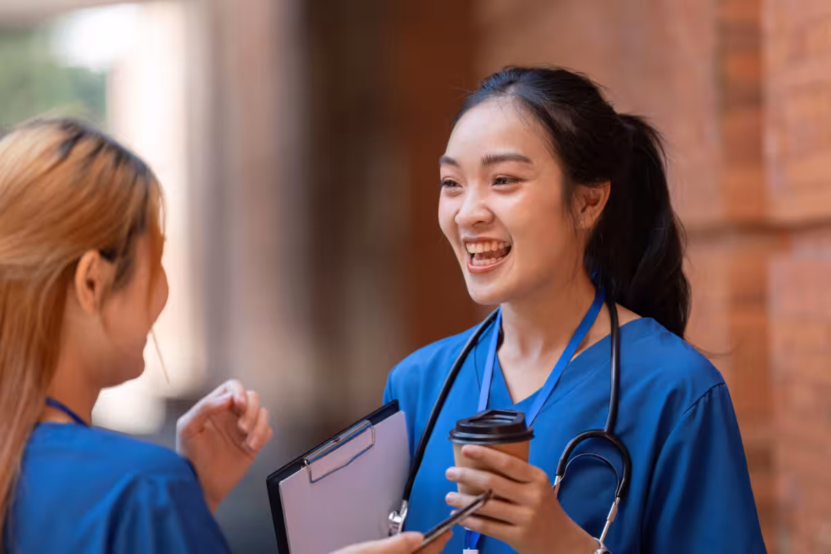 Two Nurses Blue Scrubs One Holding Coffee Cup Two nurses in blue scrubs, one holding a coffee cup, laugh together outside a building.