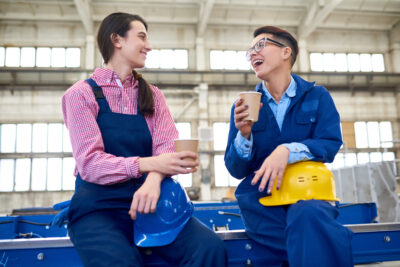 Cheerful machine operators wearing overalls chatting with each other and enjoying coffee while taking a short break from work.