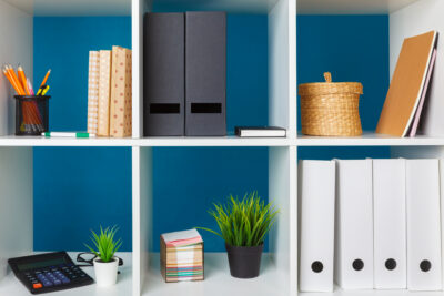 White cube shelves with office supplies including binders, books, a calculator, and plants against a blue wall.