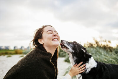 A woman laughs as a black and white dog licks her face on a beach with tall grass.