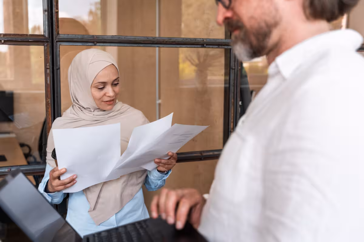 Woman Wearing Hijab Looks Papers While Man White A woman wearing a hijab looks at papers while a man in a white shirt works on a laptop.