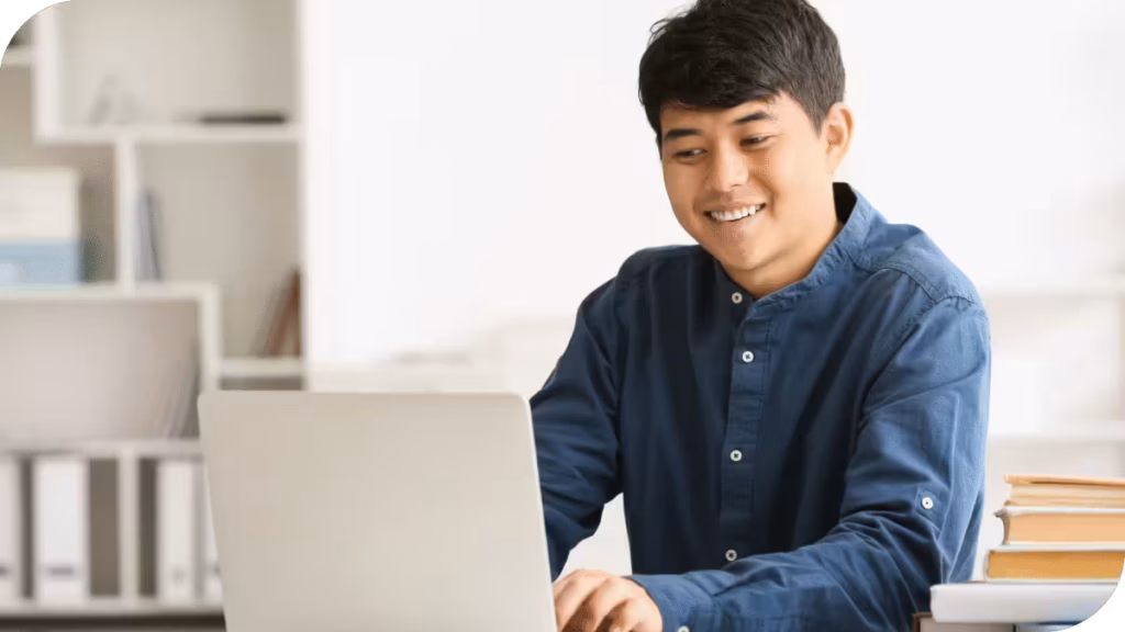 Young Asian man smiles while working on a laptop in a bright office with bookshelves in the background.