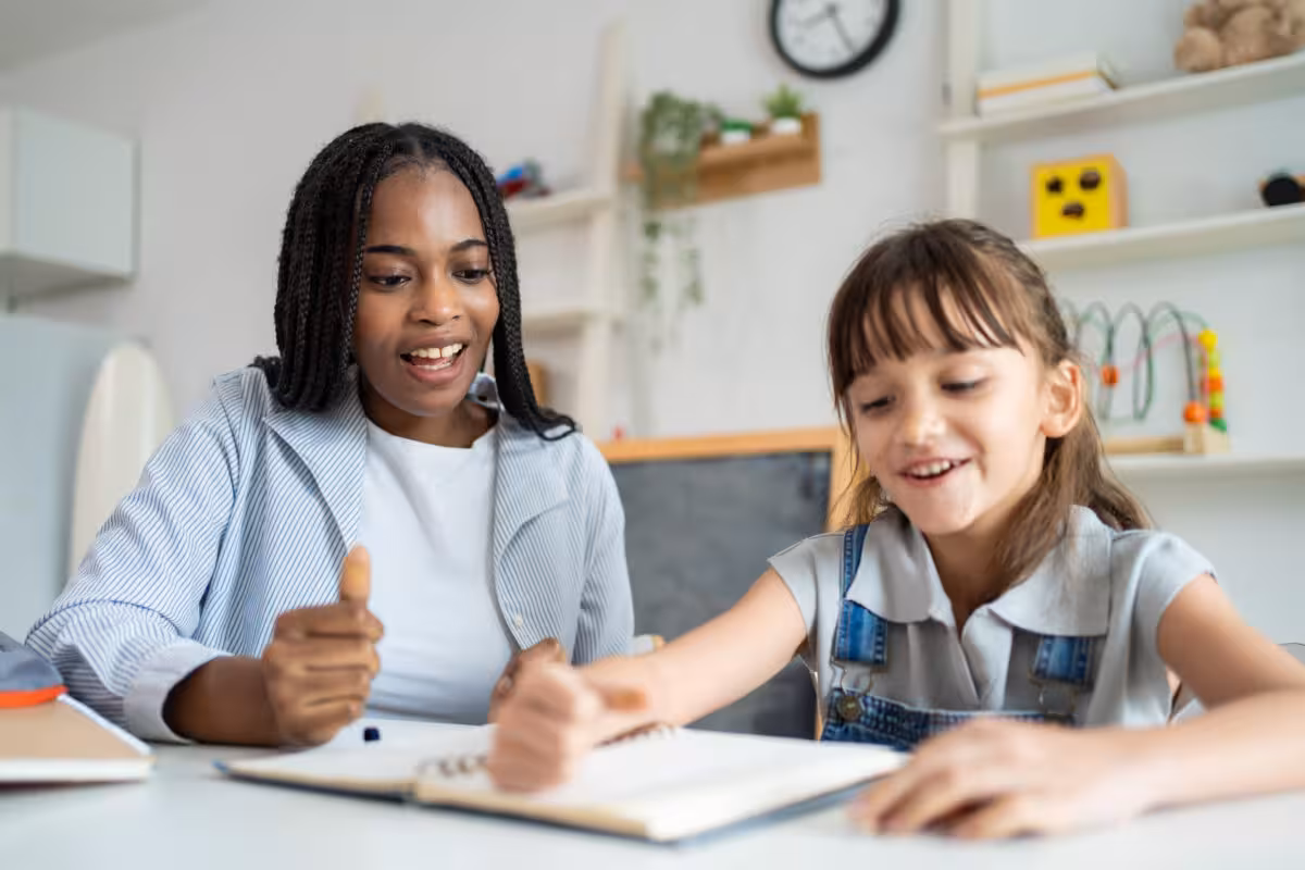 A young woman with braided hair smiles and gives a thumbs up while tutoring a happy young girl at a table.